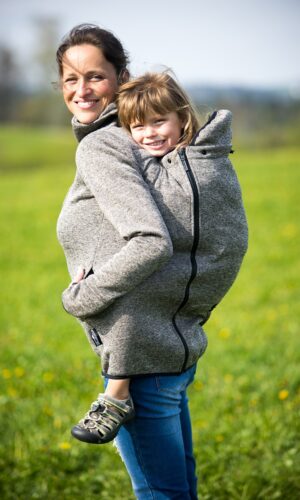 maminka nese na zádech v nosítku větší holčičku, obě se usmívají