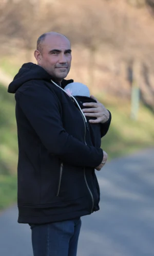 Nosící mikina Angel Wings v černé barvě na tatínkovi se spícím miminkem v nosítku, venkovní prostředí.