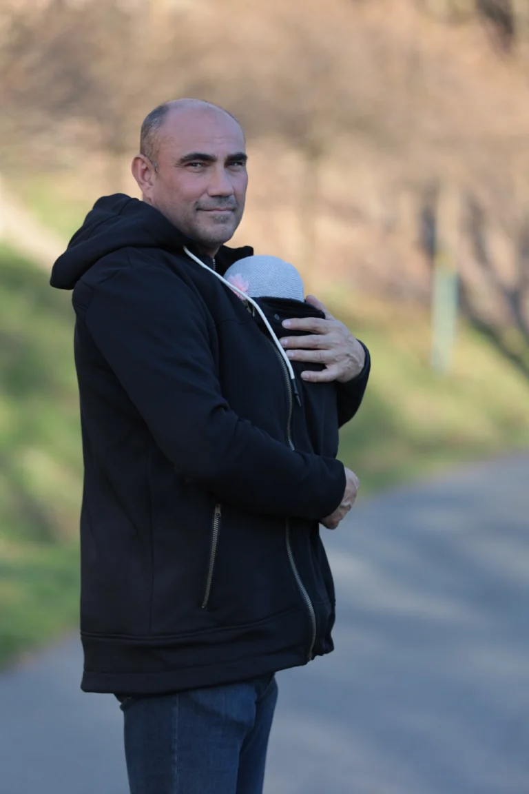 Nosící mikina Angel Wings v černé barvě na tatínkovi se spícím miminkem v nosítku, venkovní prostředí.